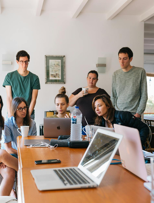 Eine Gruppe von sieben Personen sitzt und steht um einen Tisch mit Laptops und Getränken, während sie konzentriert auf einen Laptopbildschirm schauen. Der Raum ist hell und modern, mit großen Fenstern im Hintergrund.