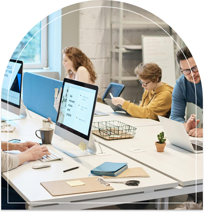 Eine Gruppe von Menschen arbeitet an einem modernen, hellen Arbeitsplatz. Mehrere Rechner sind sichtbar, und auf einem Bildschirm sind Texte über Diagramme und Grafiken zu sehen. Die Personen sind mit verschiedenen Aufgaben beschäftigt, während auf dem Tisch Schreibutensilien und Notizblöcke liegen.