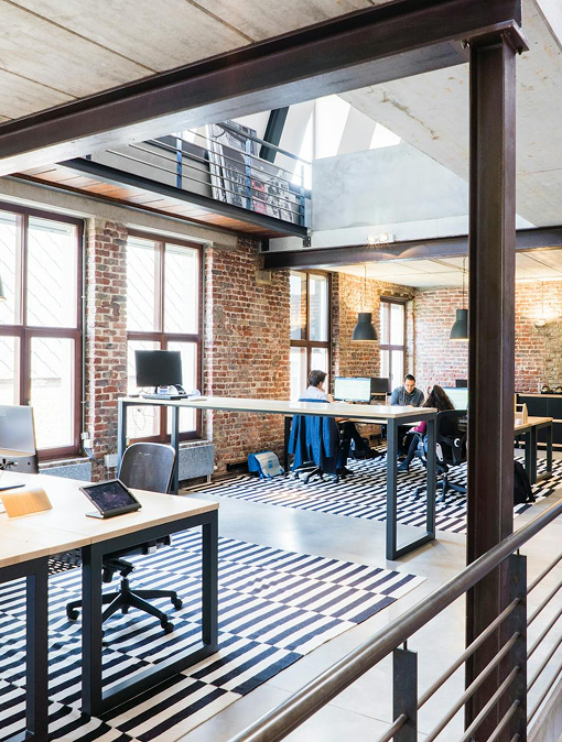 Modernes Büro mit roten Backsteinwänden und großen Fenstern. Mehrere Arbeitsplätze sind mit Computern ausgestattet, und es sind Personen an Tischen zu sehen. Der Boden ist mit einem gestreiften Teppich dekoriert.