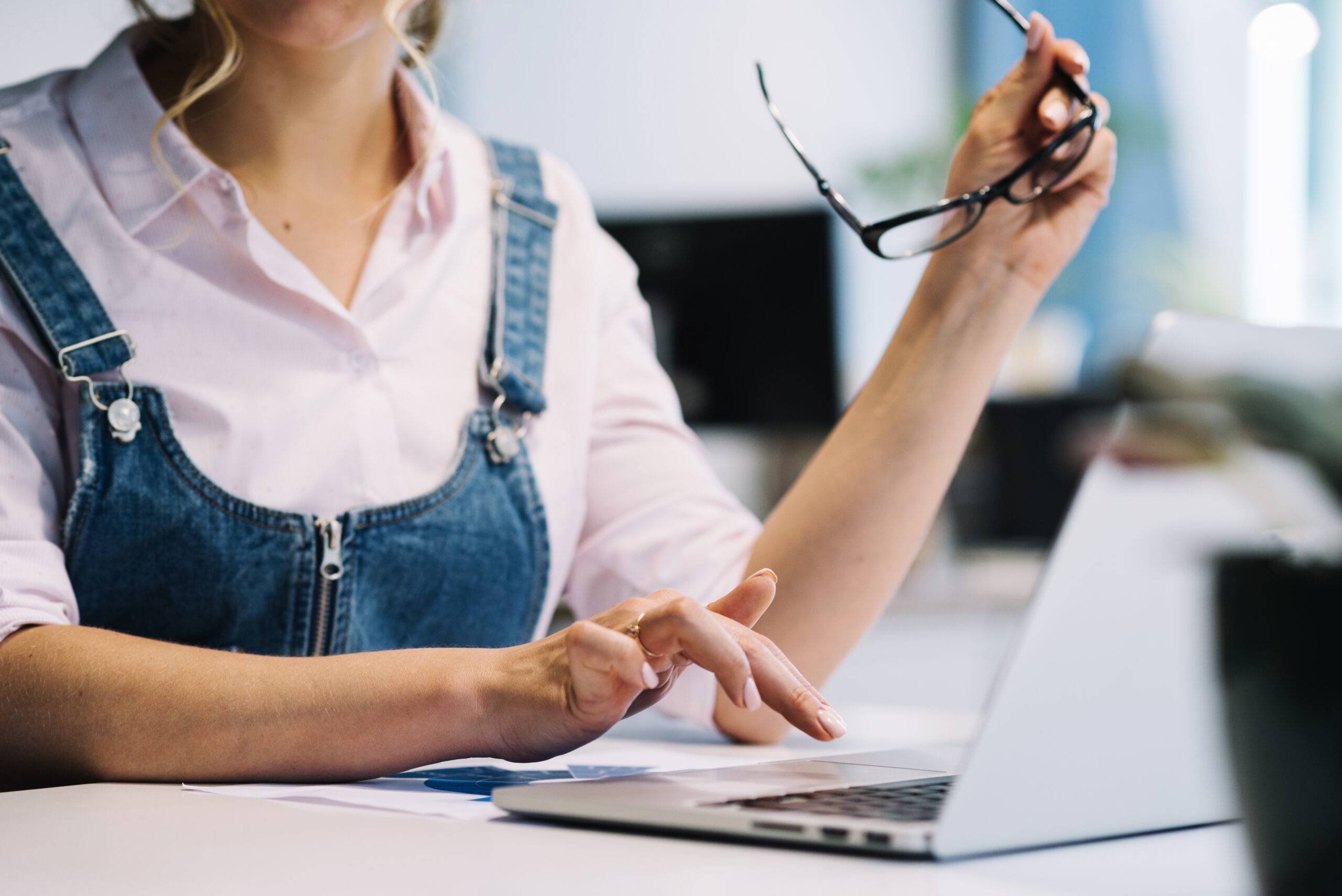 Eine Person mit einem rosa Hemd und einer Jeans-Latzhose sitzt an einem Tisch und arbeitet am Laptop. Sie hält eine Brille in der rechten Hand und tippt mit der linken Hand auf die Tastatur. Im Hintergrund sind unscharfe Büroelemente zu sehen.