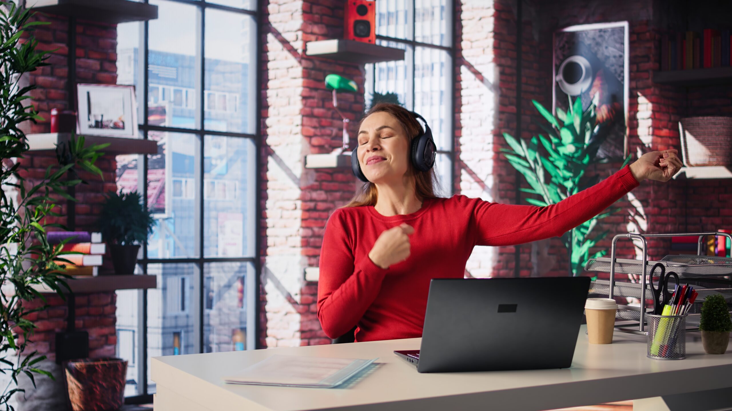 Eine Frau mit Kopfhörern trägt ein rotes Oberteil und sitzt an einem Schreibtisch mit einem Laptop. Sie scheint Musik zu genießen, während sie mit einem Arm in die Luft zeigt, vor großen Fenstern in einem modernen Raum mit Pflanzen und Regalen im Hintergrund.